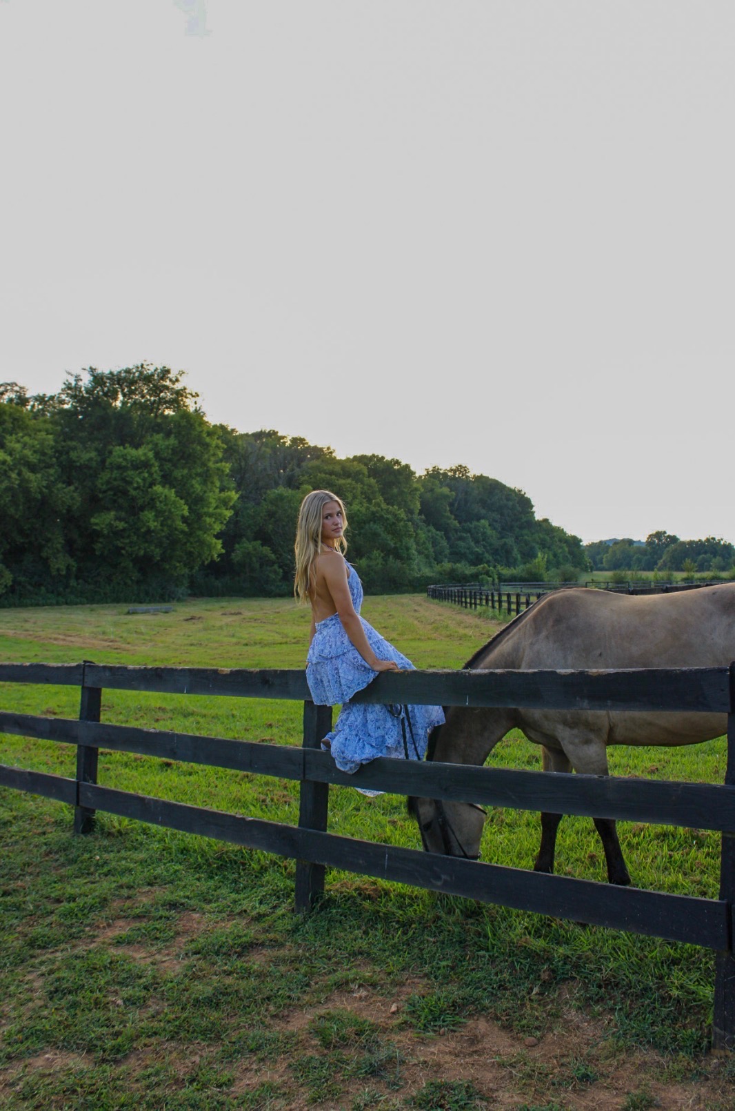 Girl seated on a black farm fence with a grazing horse